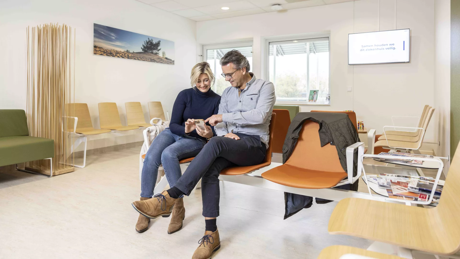 Een koppel zit in een wachtkamer en kijkt samen naar een smartphone.