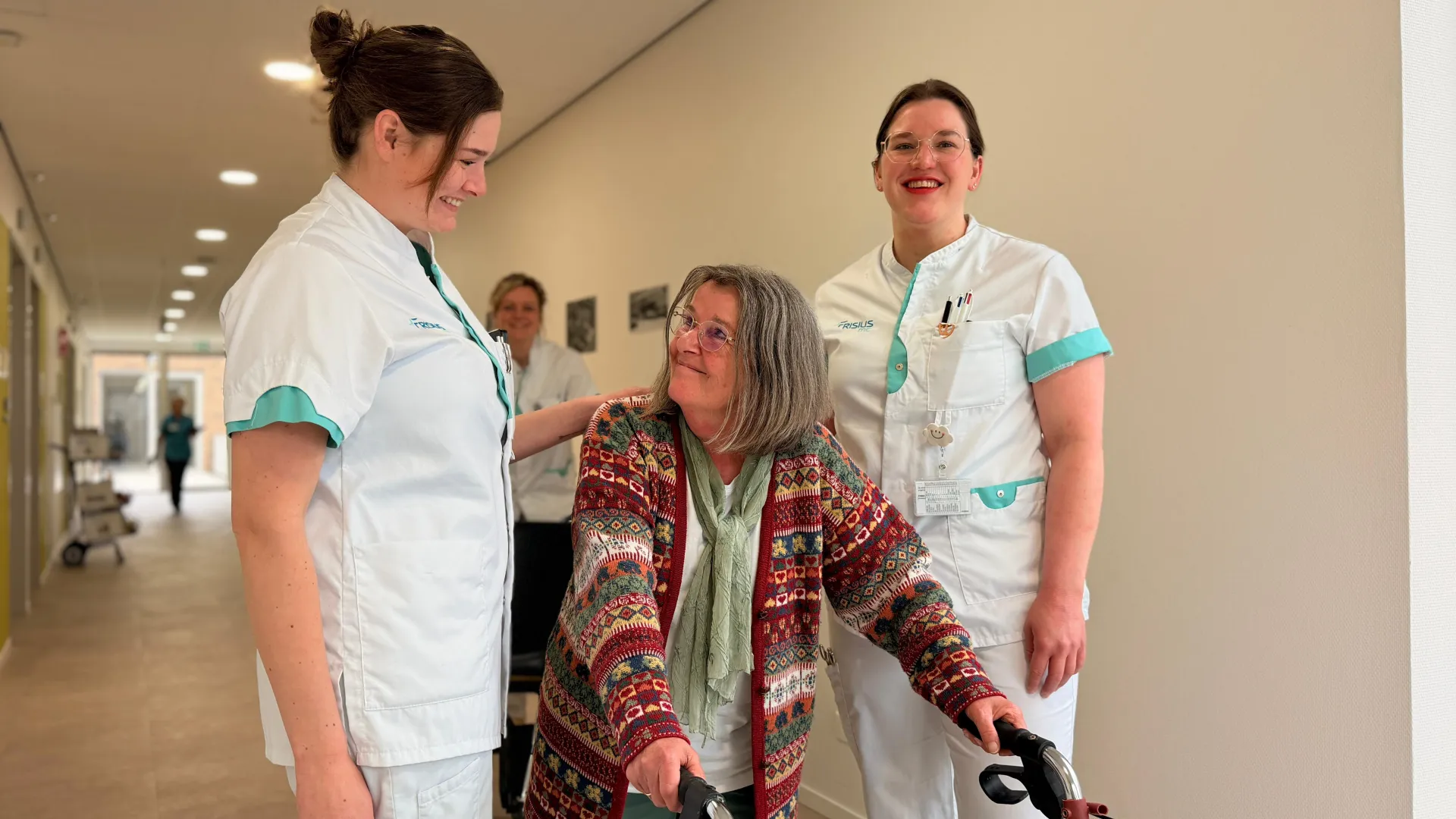 Verpleegkundigen helpen een oudere vrouw met een rollator in een gang.