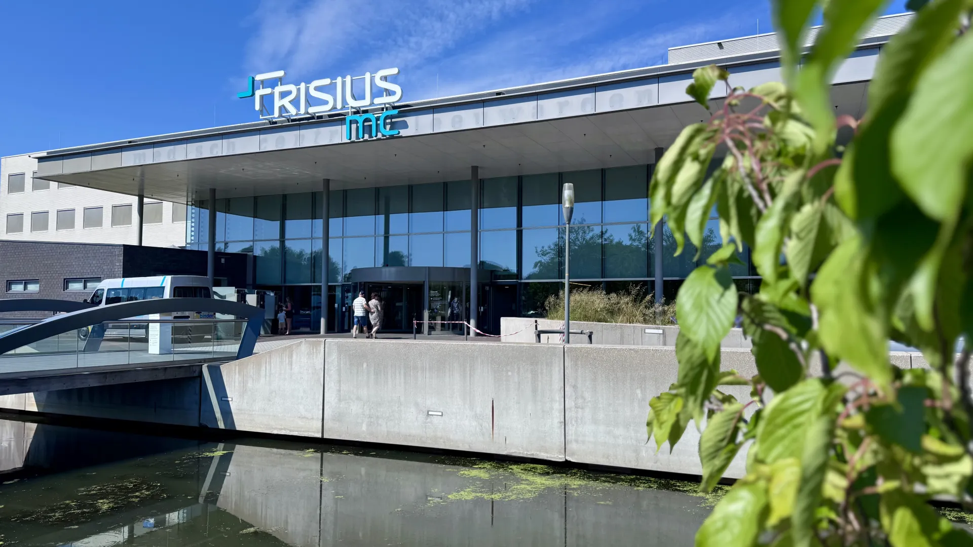 Vooraanzicht van Frisius MC Leeuwarden met een vijver op de voorgrond onder een blauwe hemel.