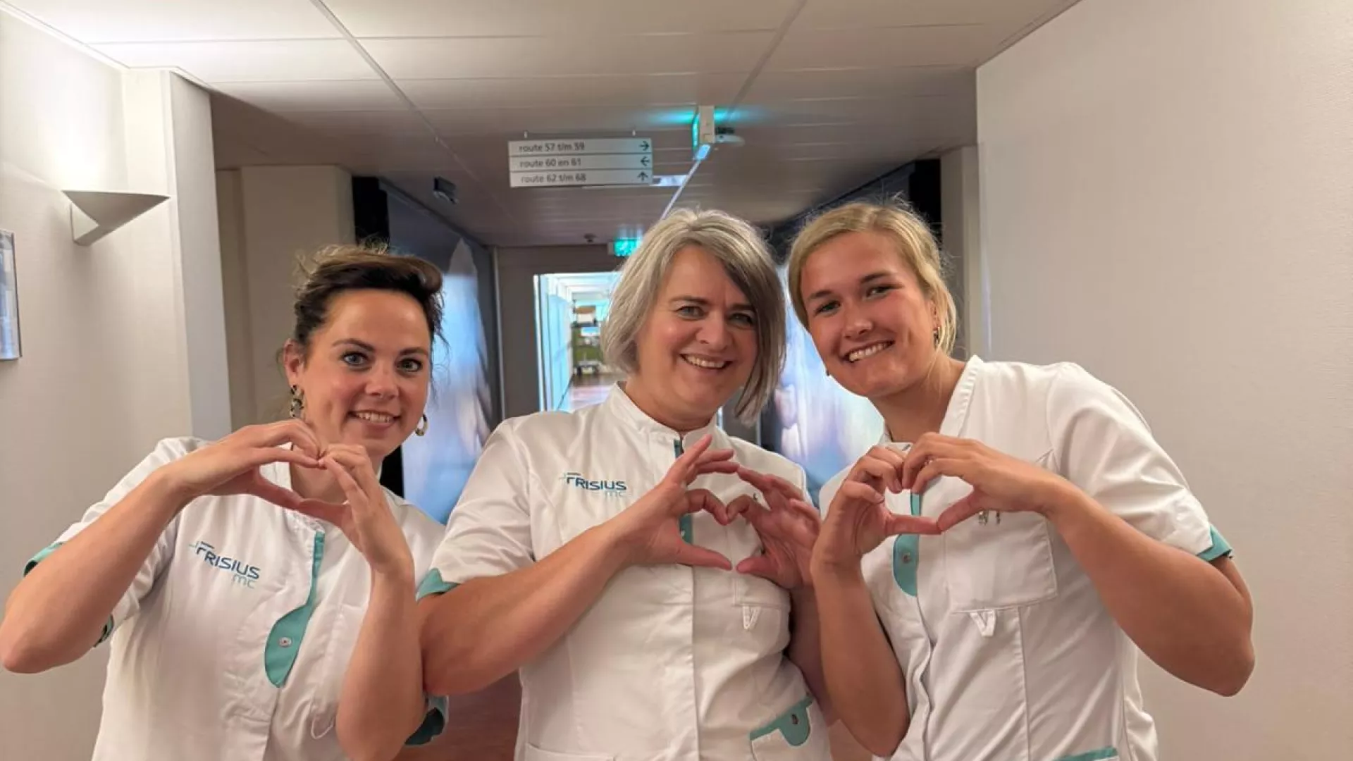 Drie verpleegkundigen vormen hartjes met hun handen in de ziekenhuisgang.