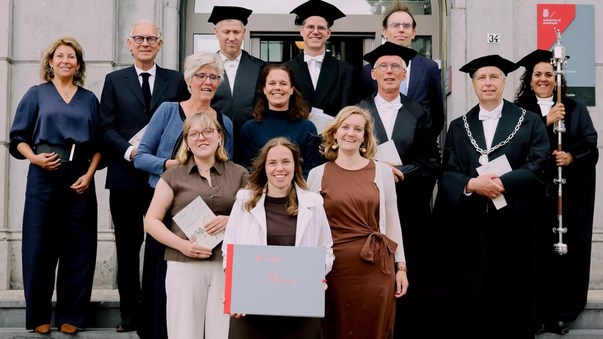 Een groep mensen staat op trappen voor een gebouw, sommigen in academische kledij.