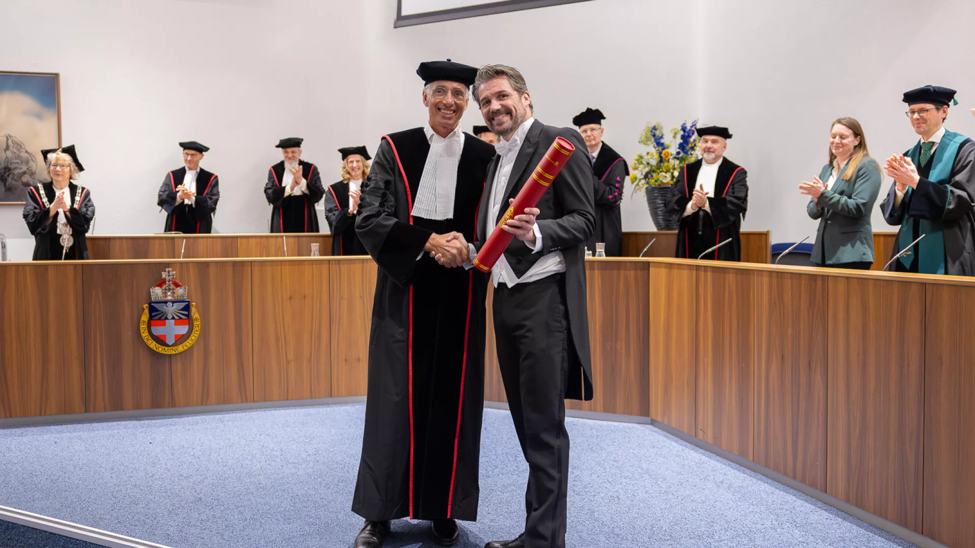 Twee mannen glimlachen en schudden handen tijdens een ceremonie, met applaudisserende mensen op de achtergrond.