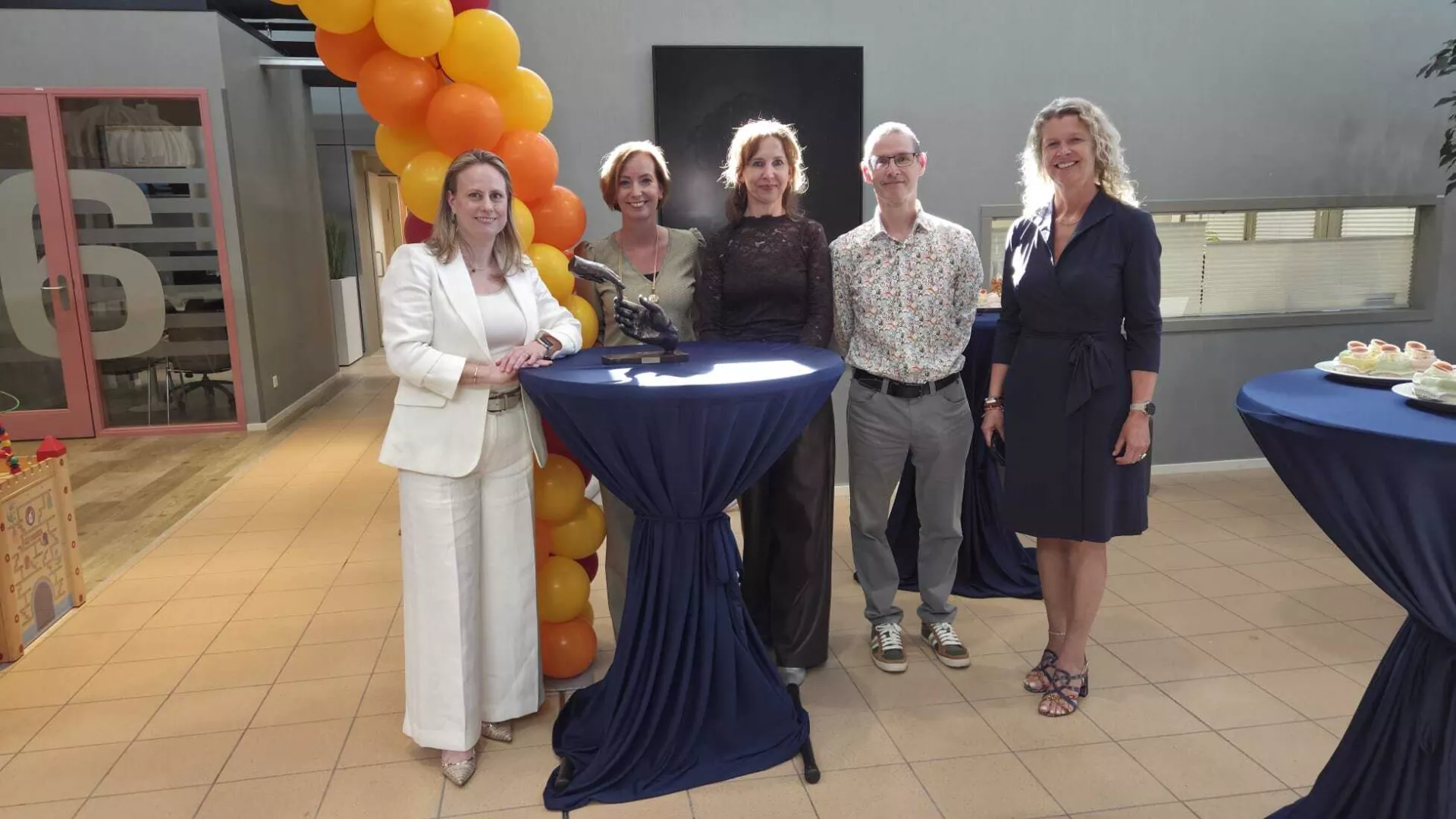 Groepsfoto van vijf mensen bij een sta-tafel met ballondecoratie op de achtergrond.
