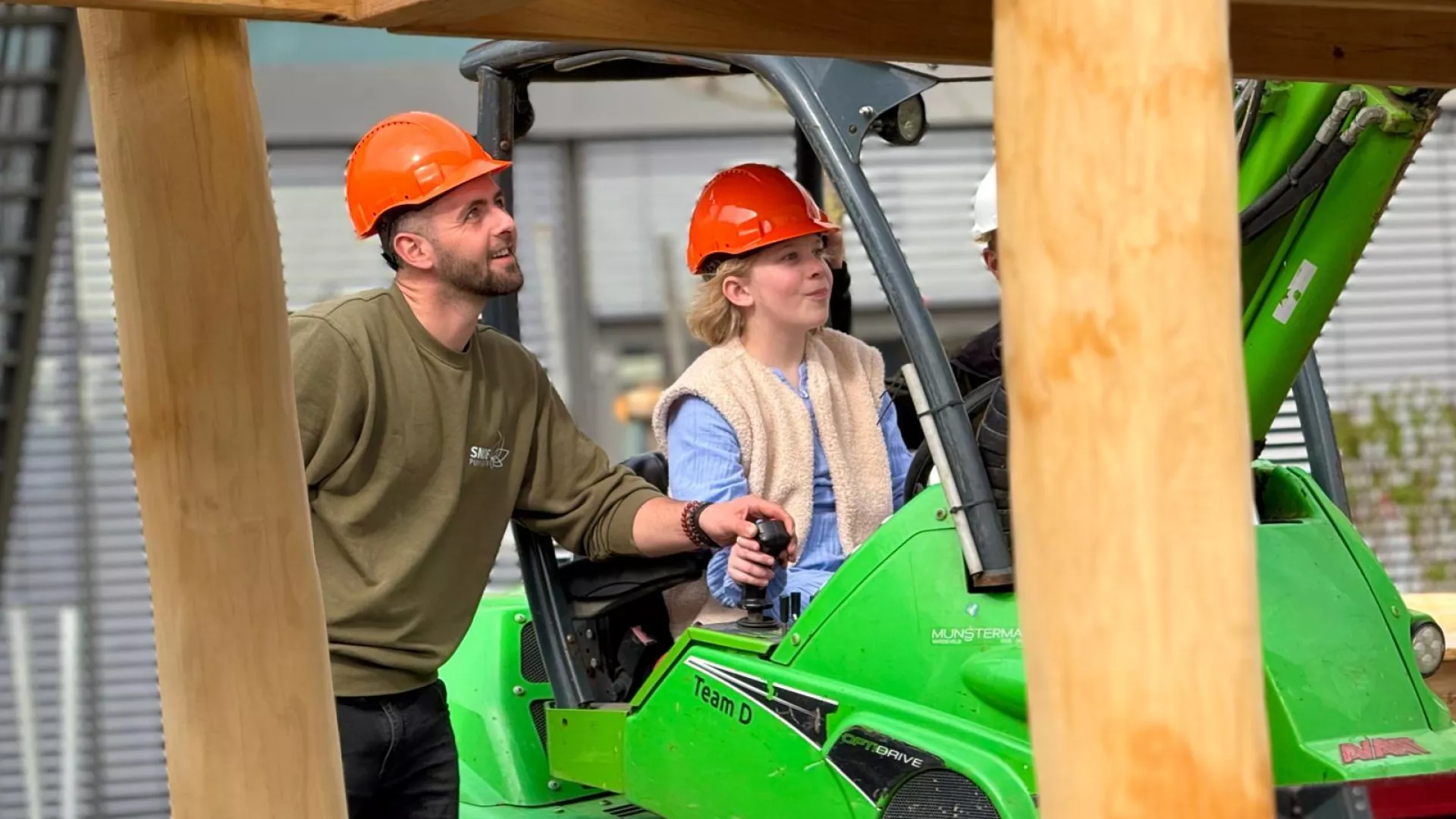 Twee personen met bouwhelmen besturen een groen voertuig op een bouwplaats.