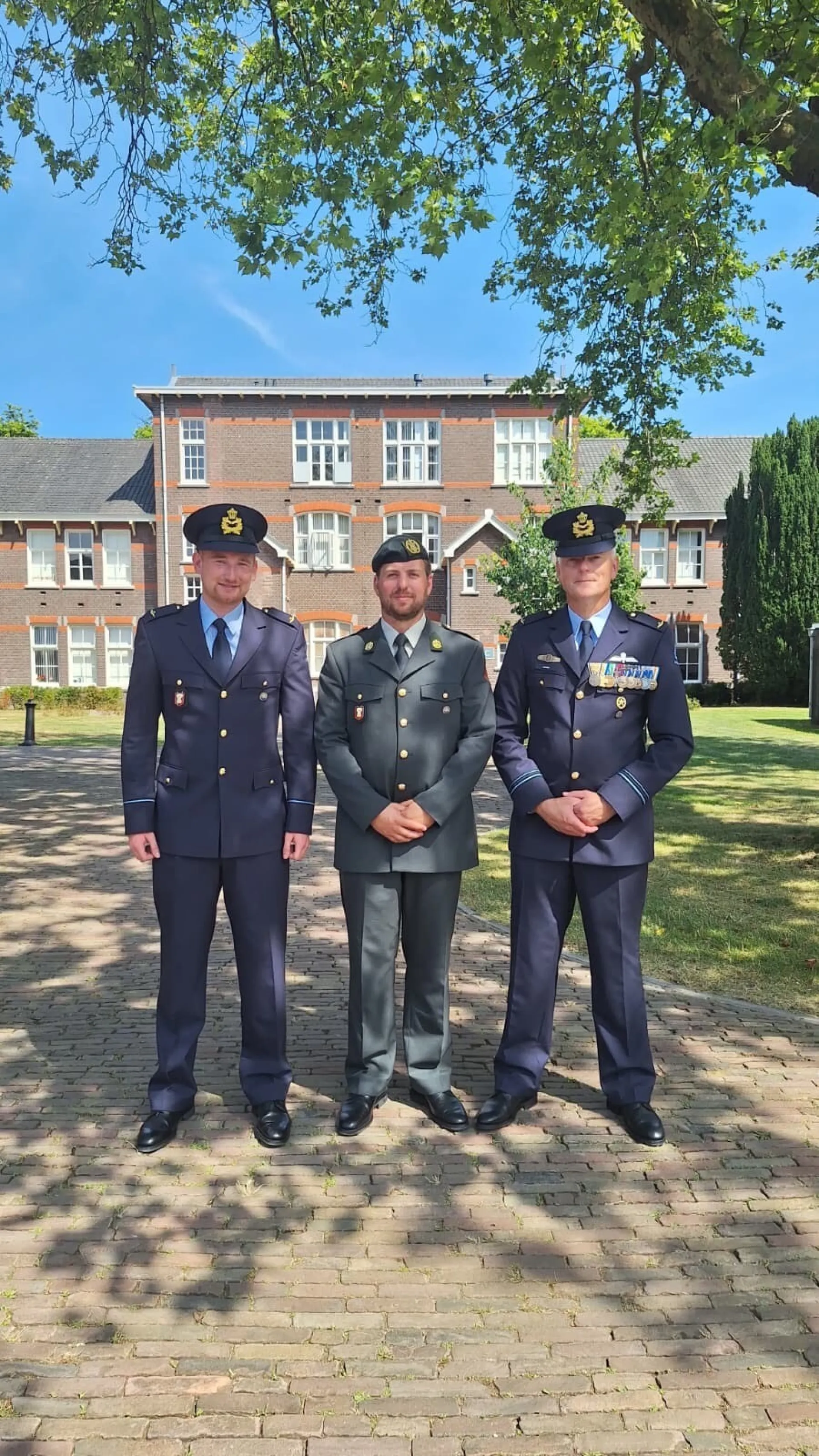 Drie mannen in militaire uniformen poseren voor een gebouw in een zonnige buitenomgeving.