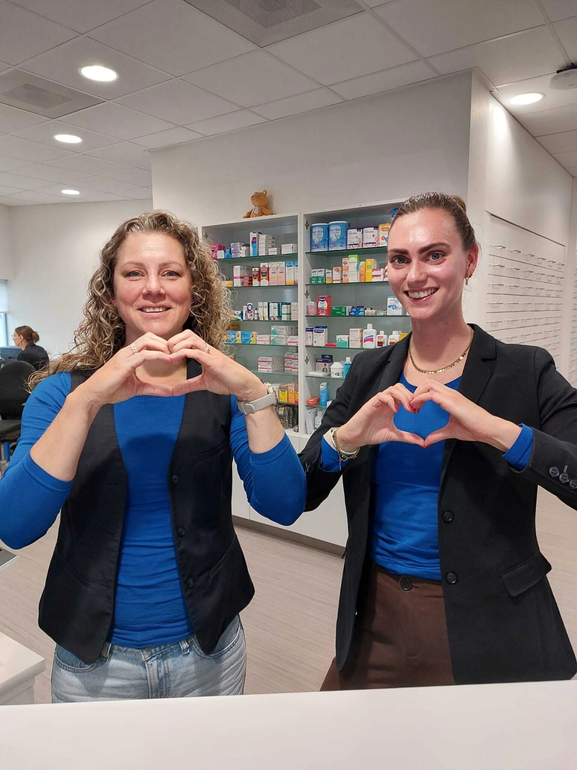 Twee vrouwen maken hartvormen met hun handen in een apotheek.