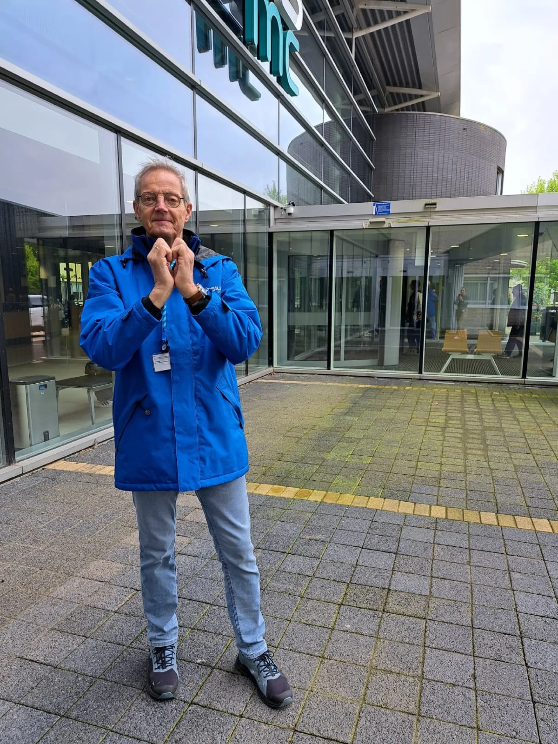 Een persoon in een blauwe jas staat voor het ziekenhuis en vormt een hartje met zijn handen.