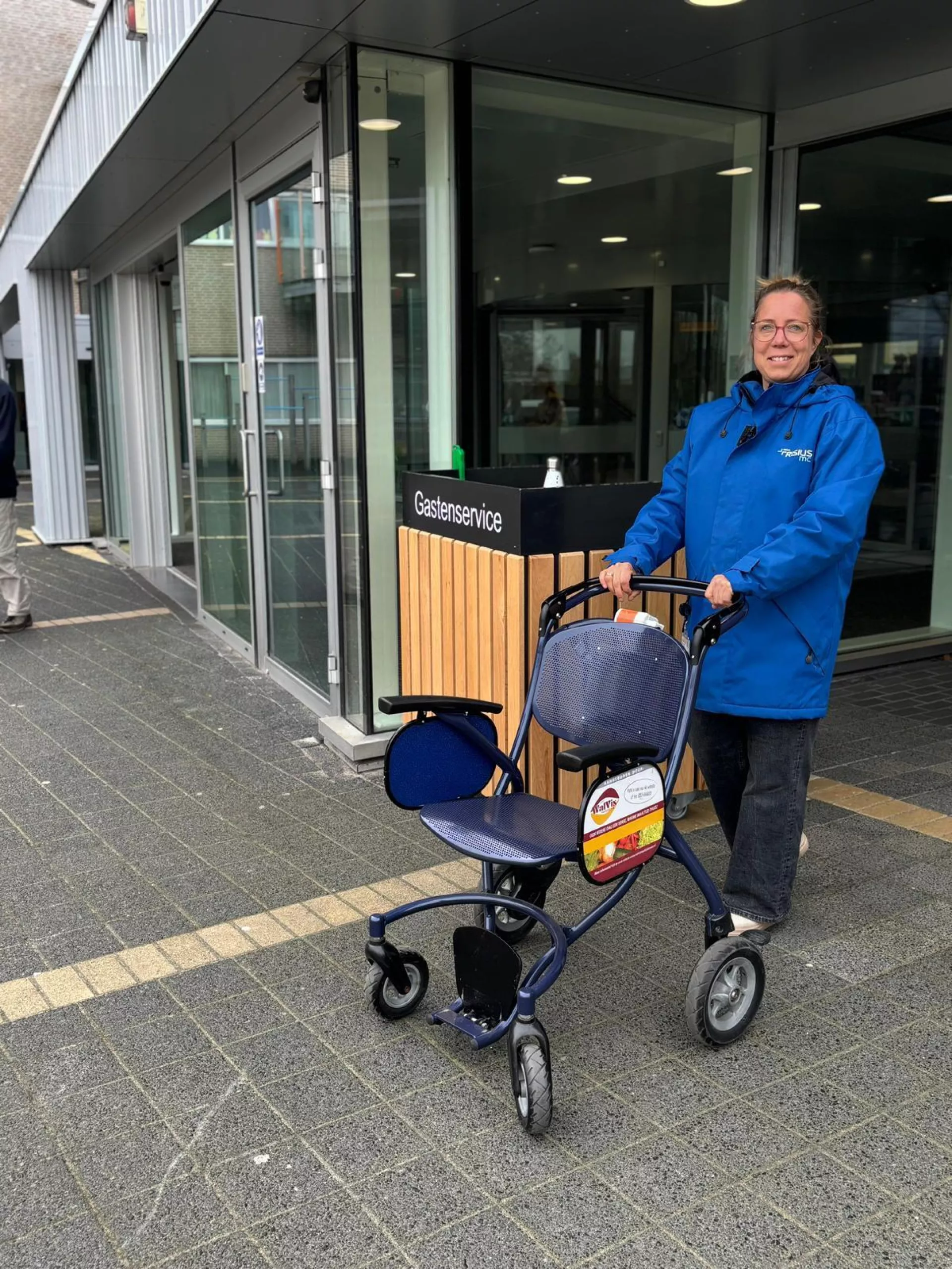 Een medewerker met een blauwe jas duwt een rollator buiten een gebouw.