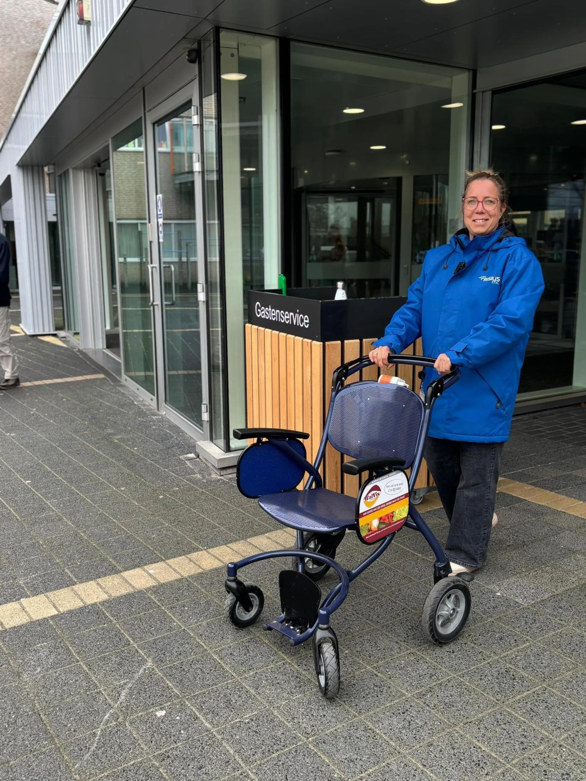 Een medewerker met een blauwe jas duwt een rollator buiten een gebouw.