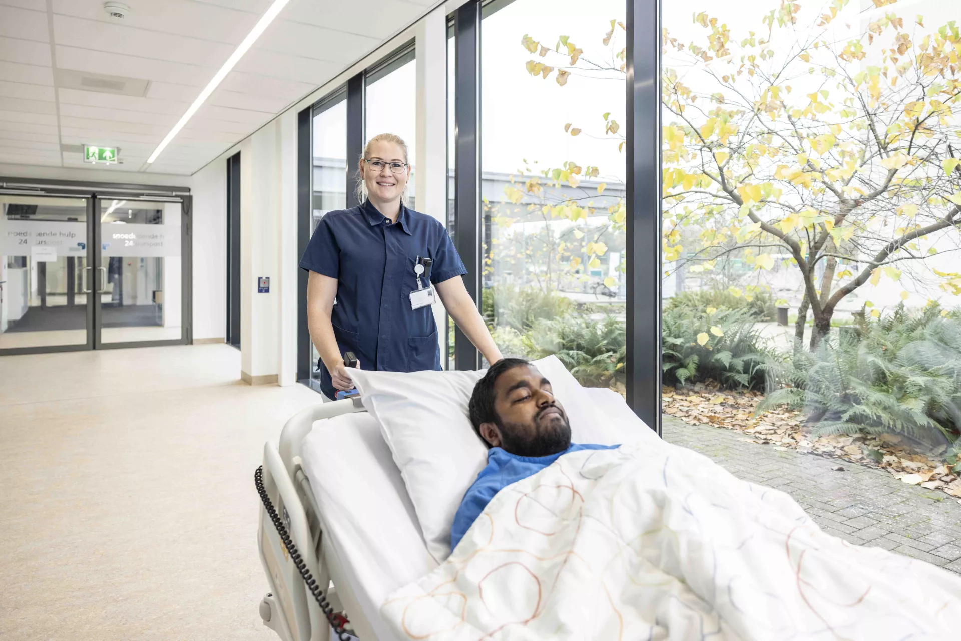 Medewerker van het ziekenhuis begeleidt een patiënt in bed door een gang.