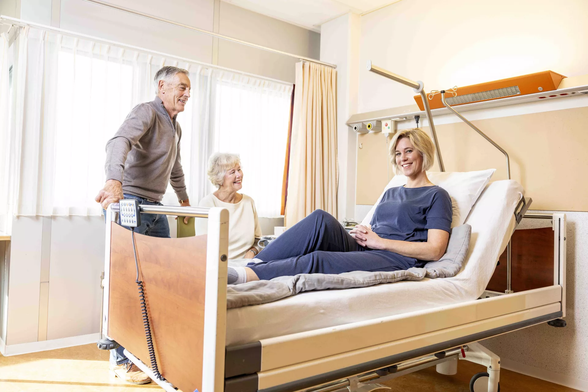 Een vrouw ligt in een ziekenhuisbed terwijl twee oudere mensen aan haar zijde staan en glimlachen.