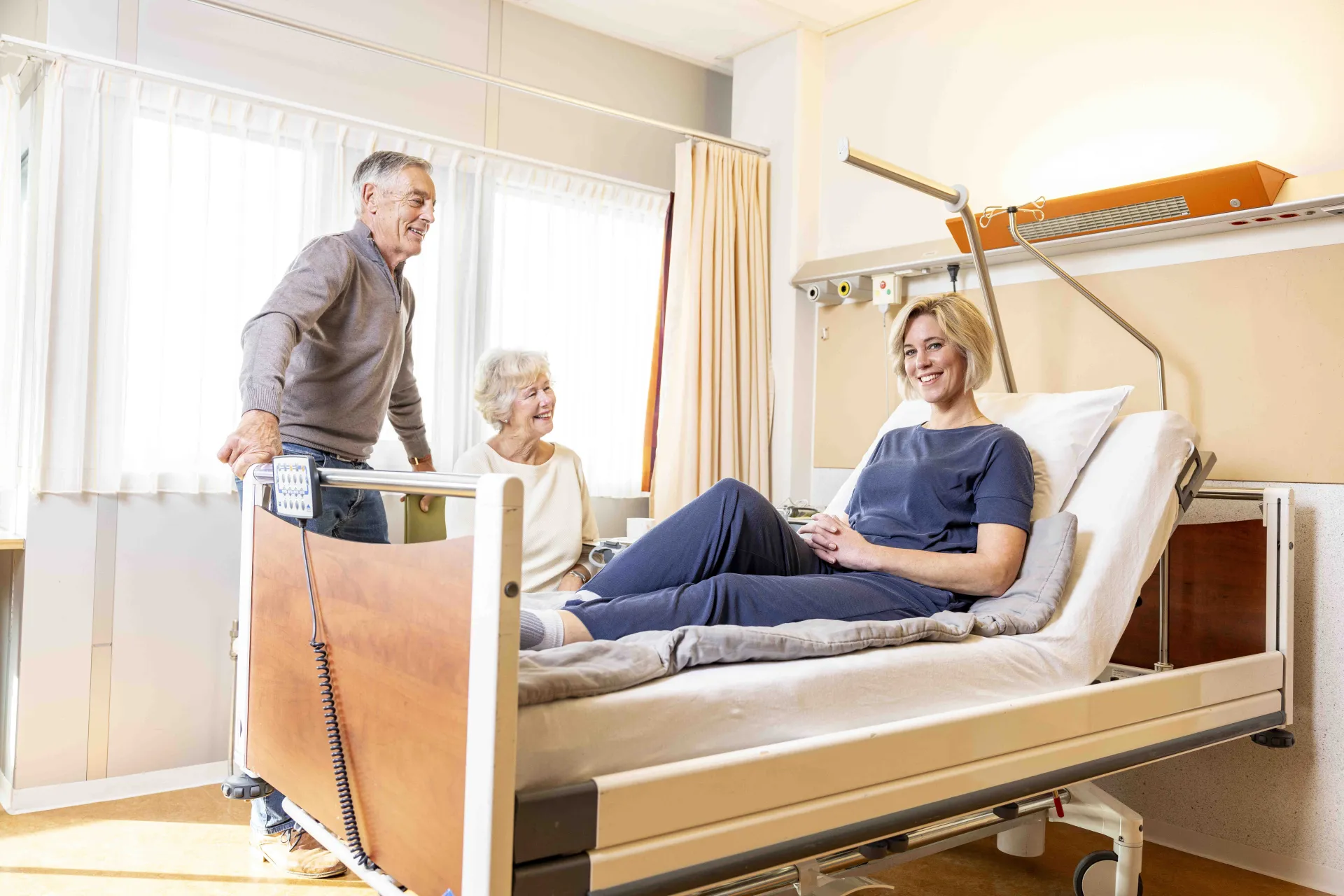 Een vrouw ligt in een ziekenhuisbed terwijl twee oudere mensen aan haar zijde staan en glimlachen.