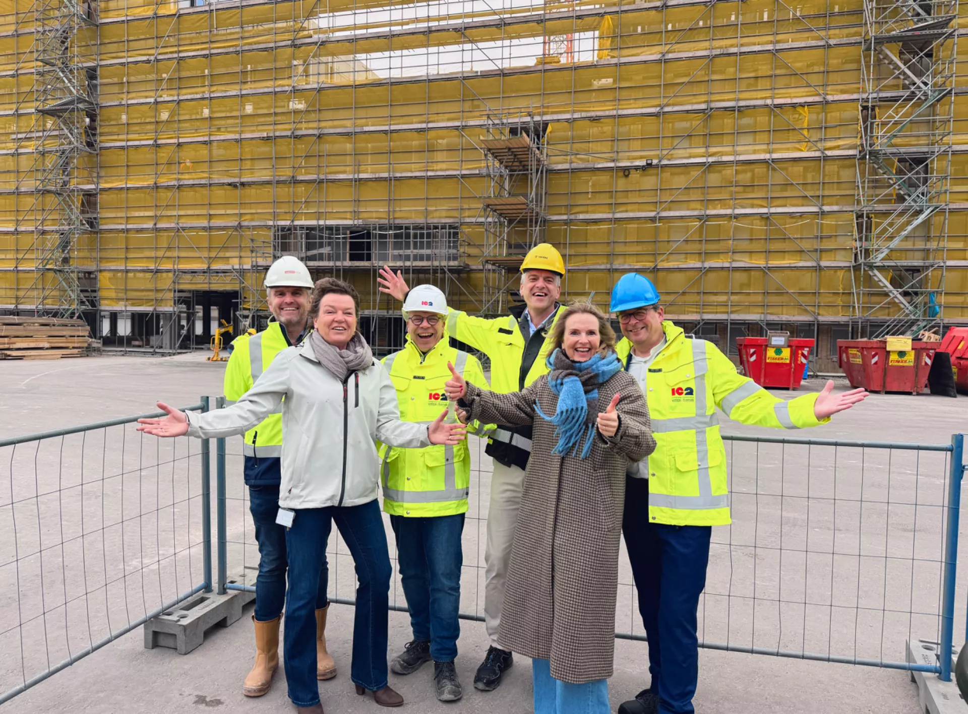 Groep mensen poseert met plezier op een bouwplaats, gekleed in veiligheidskleding en helmen.