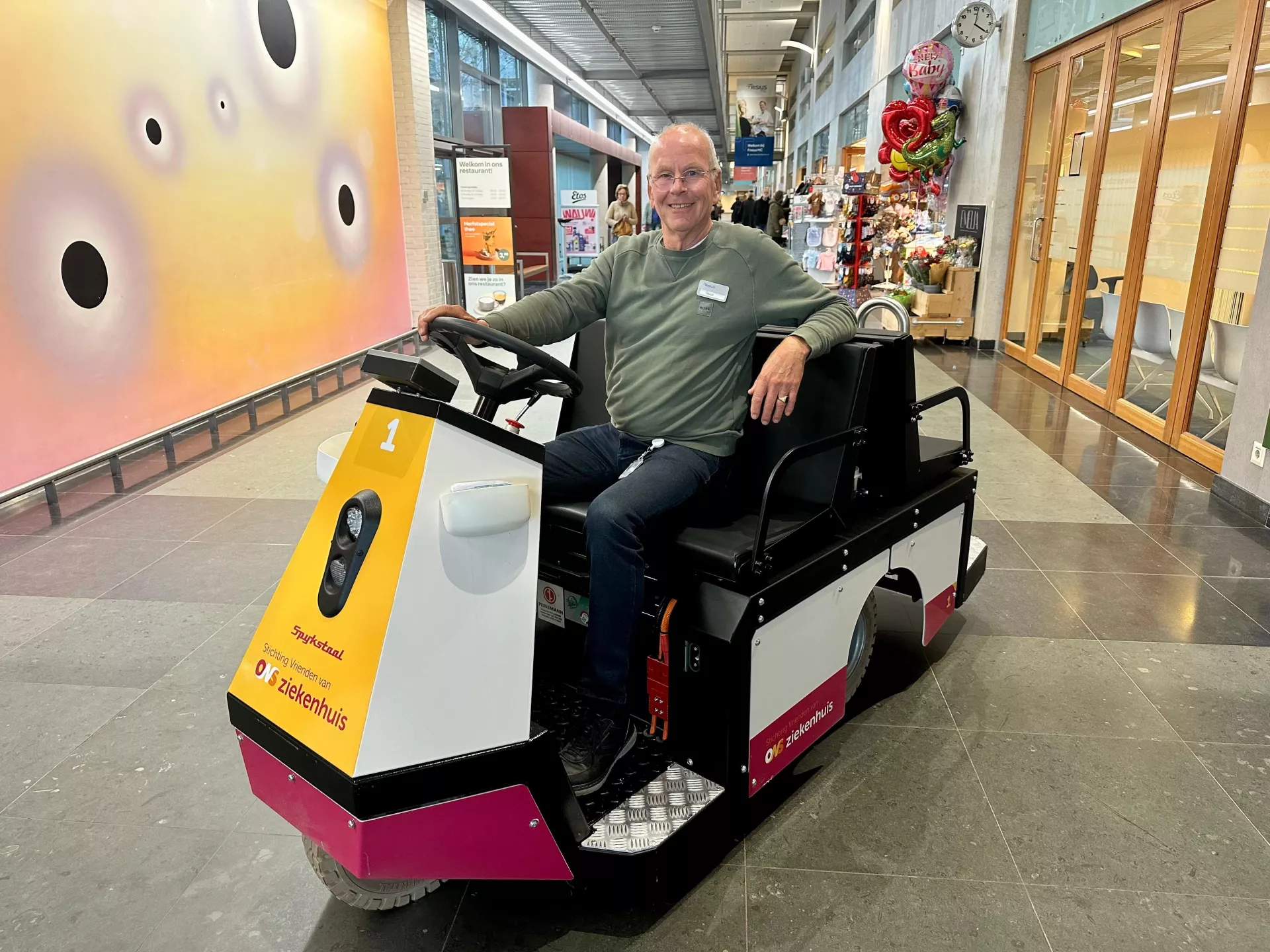Een man zit lachend op een kleine elektrische kar in de centrale hal van het ziekenhuis.