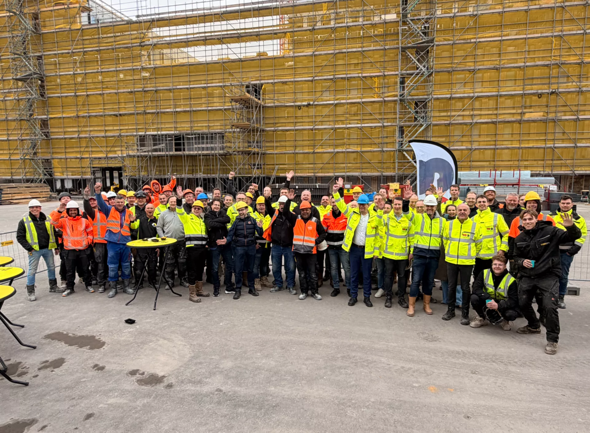 Een groep bouwvakkers poseert voor een constructieplaats met steigers.