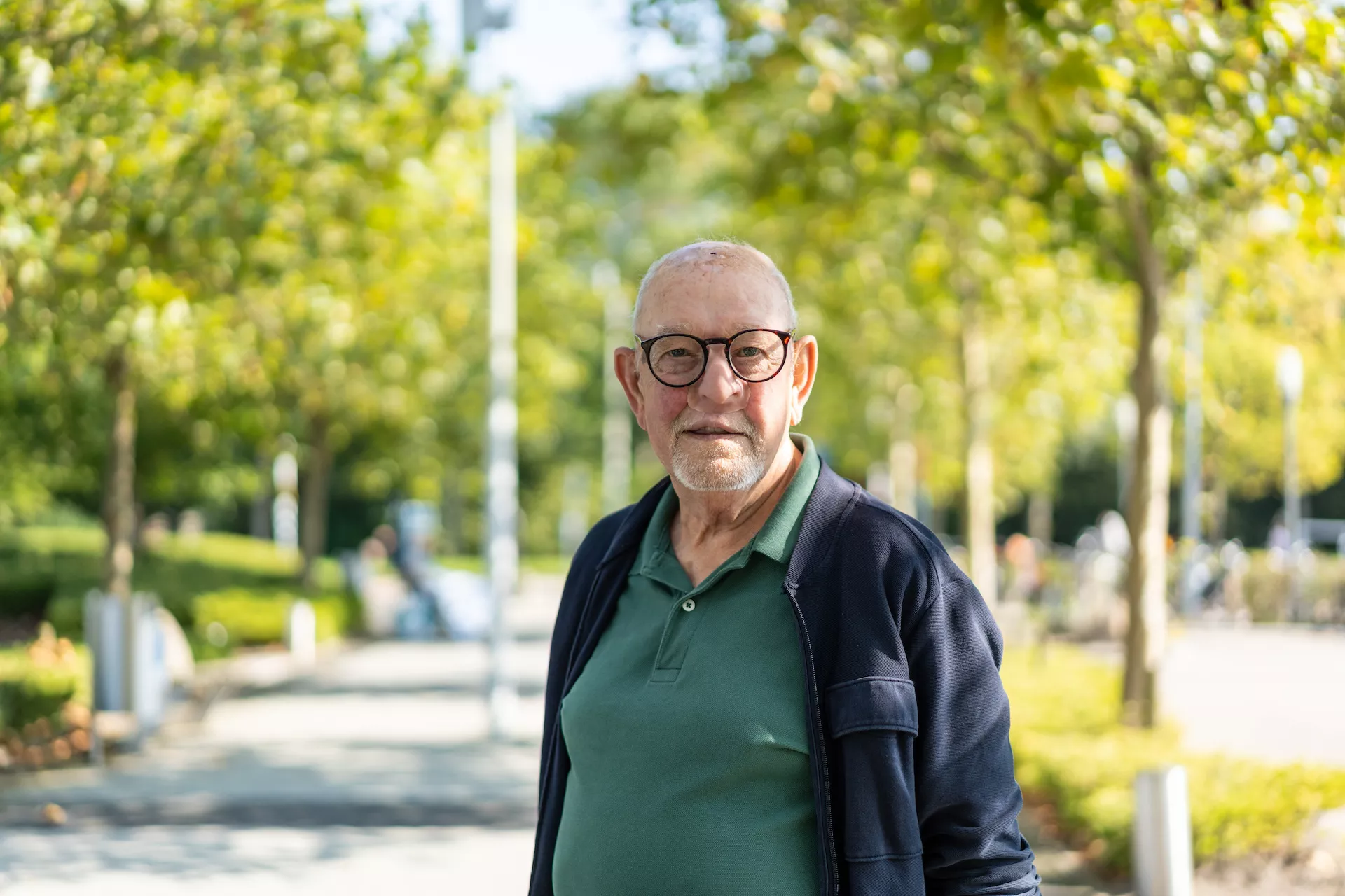 Een oudere man poseert in een zonnig park met bomen op de achtergrond.