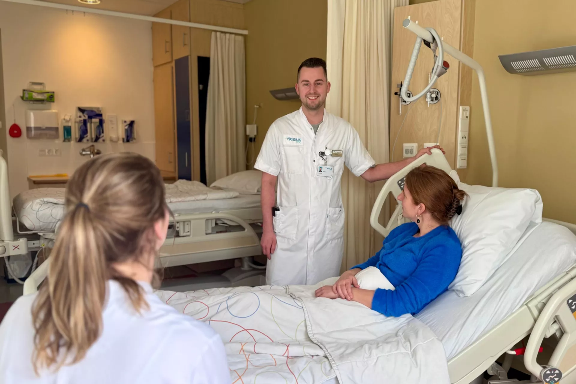 Een verpleegkundige spreekt met een patiënt in een ziekenhuisbed, terwijl een andere persoon toekijkt.