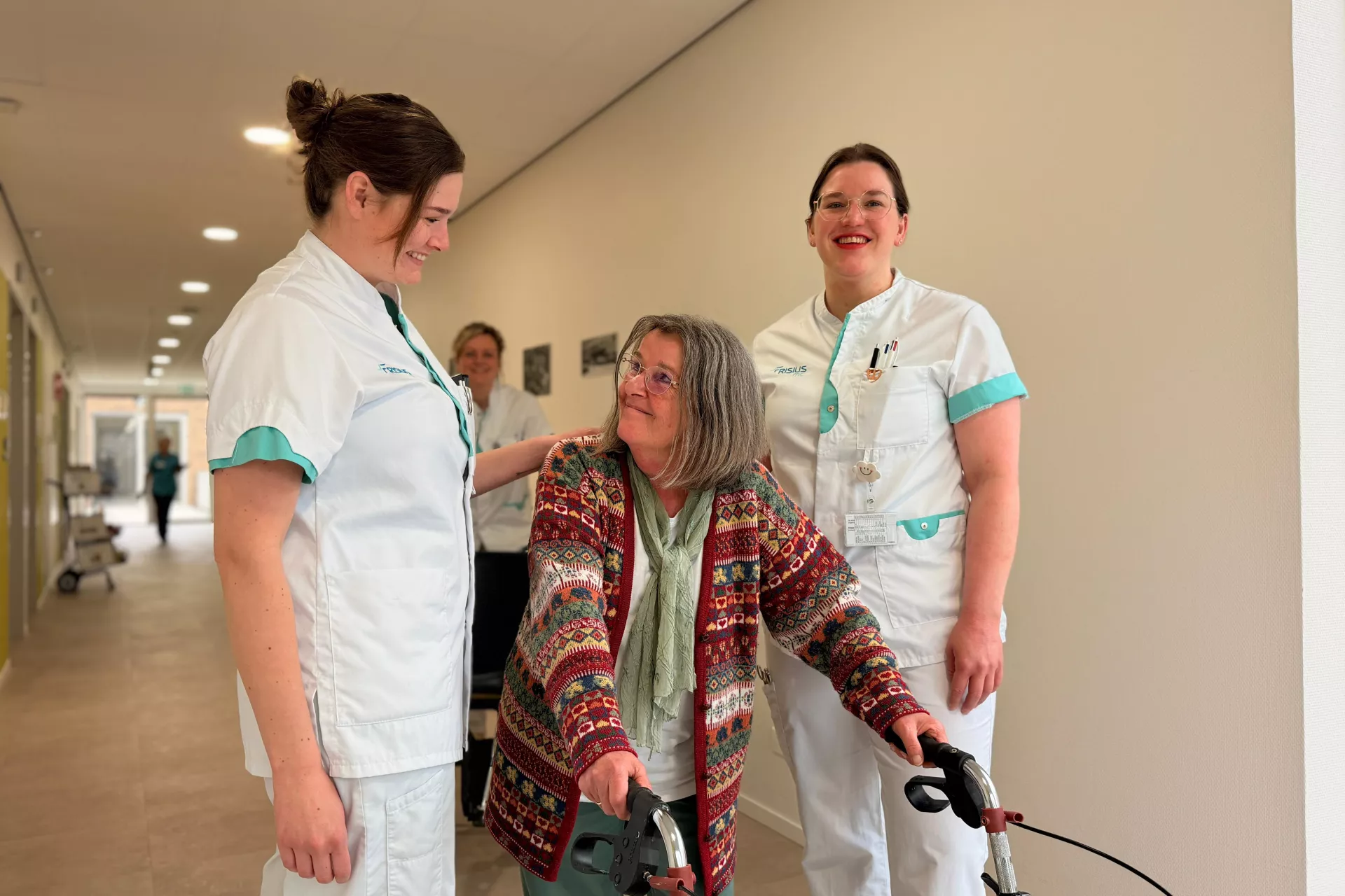Verpleegkundigen helpen een oudere vrouw met een rollator in een gang.