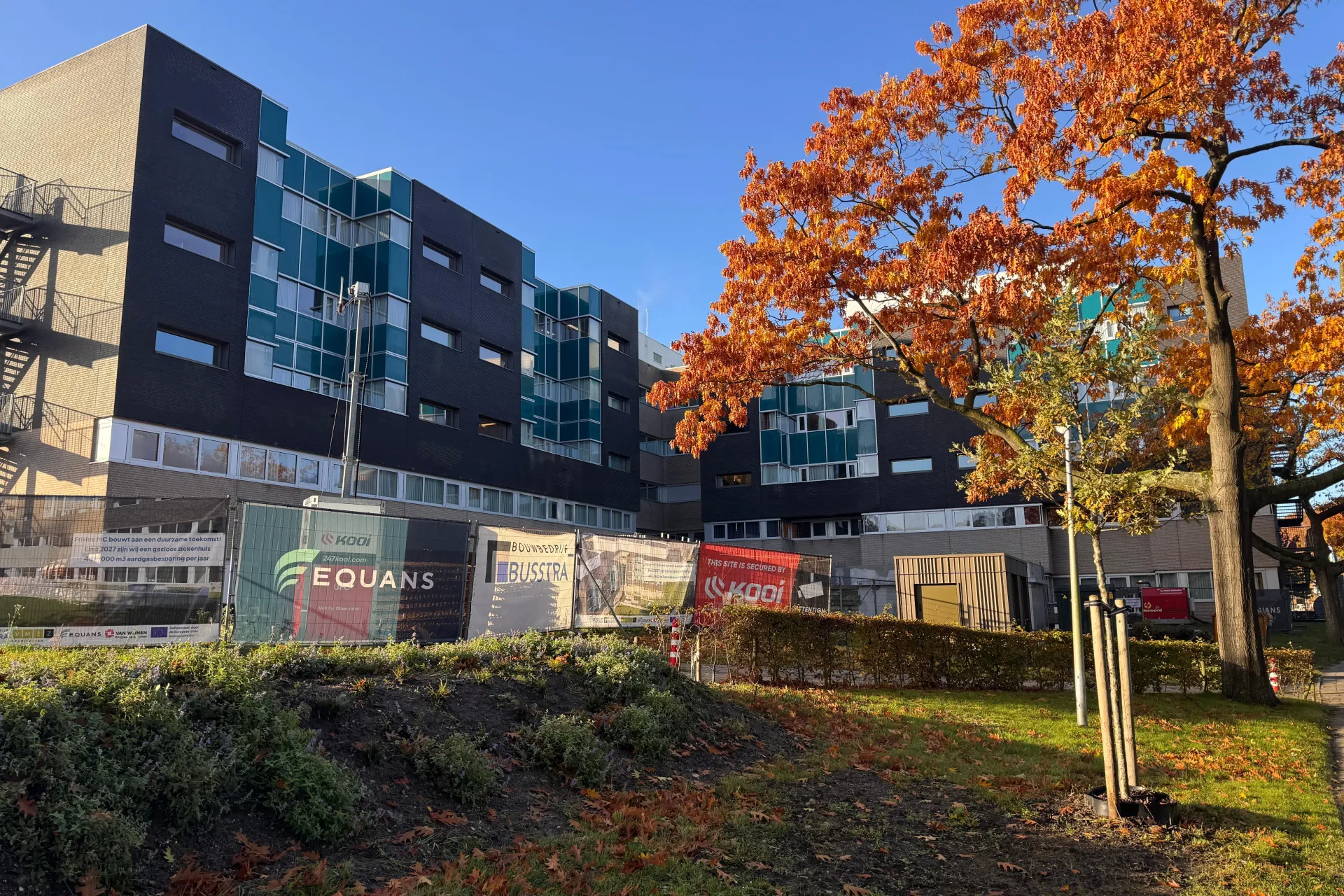 Bouwplaats naast beddenhuis van Frisius MC Heerenveen in de herfstzon met kleurrijke bomen op de voorgrond.