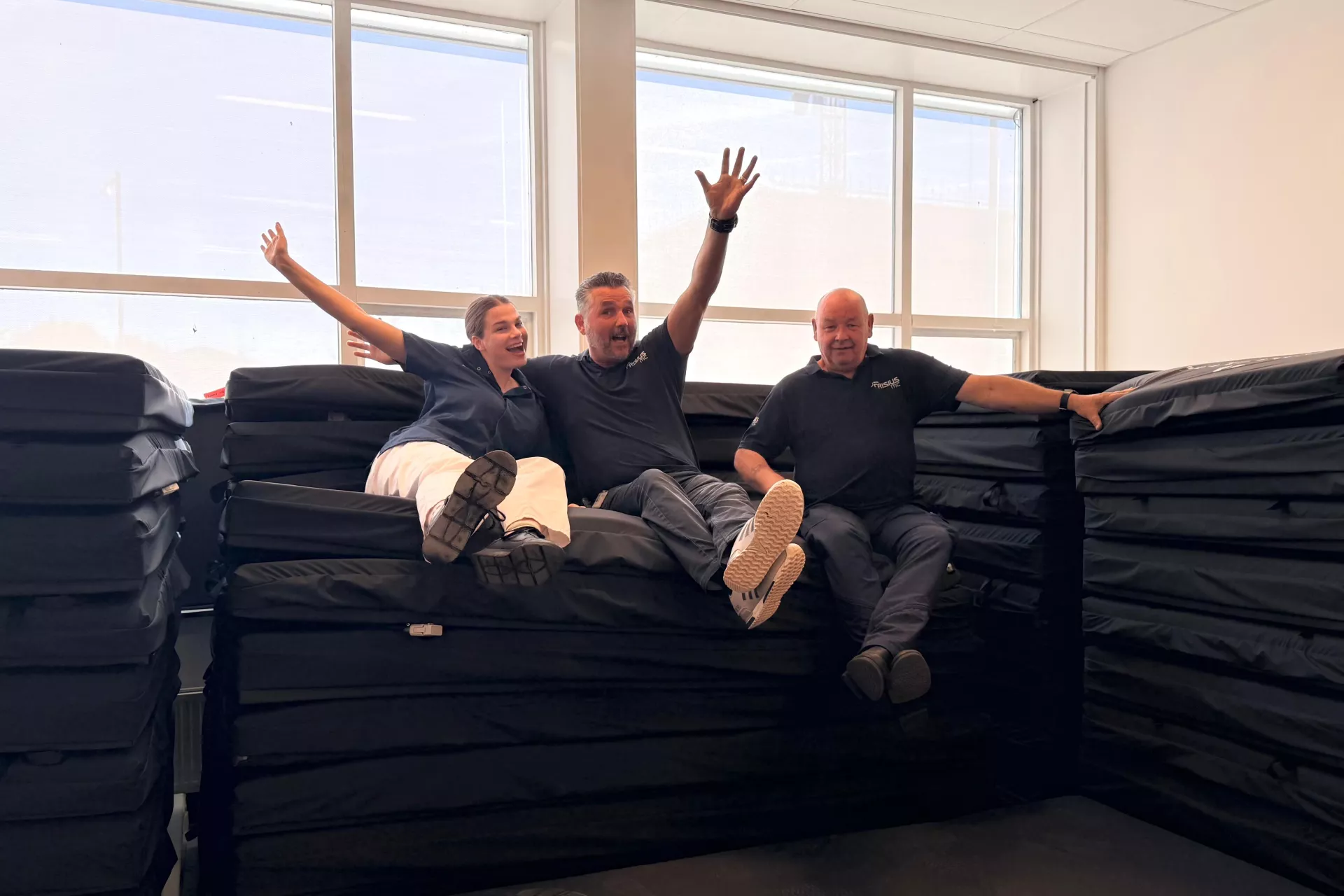 Drie mensen zitten vrolijk op een stapel matrassen in een kamer.