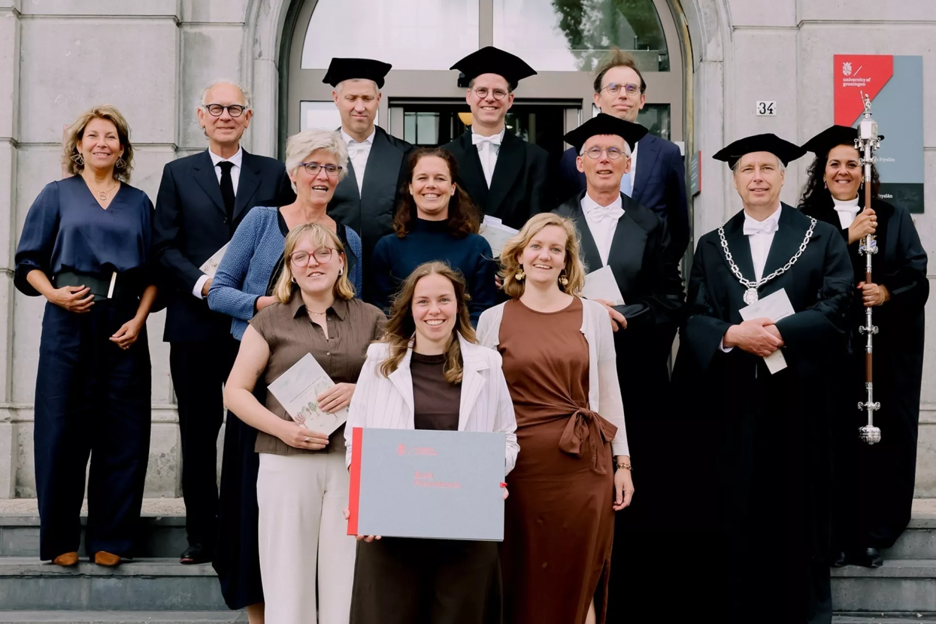 Een groep mensen staat op trappen voor een gebouw, sommigen in academische kledij.