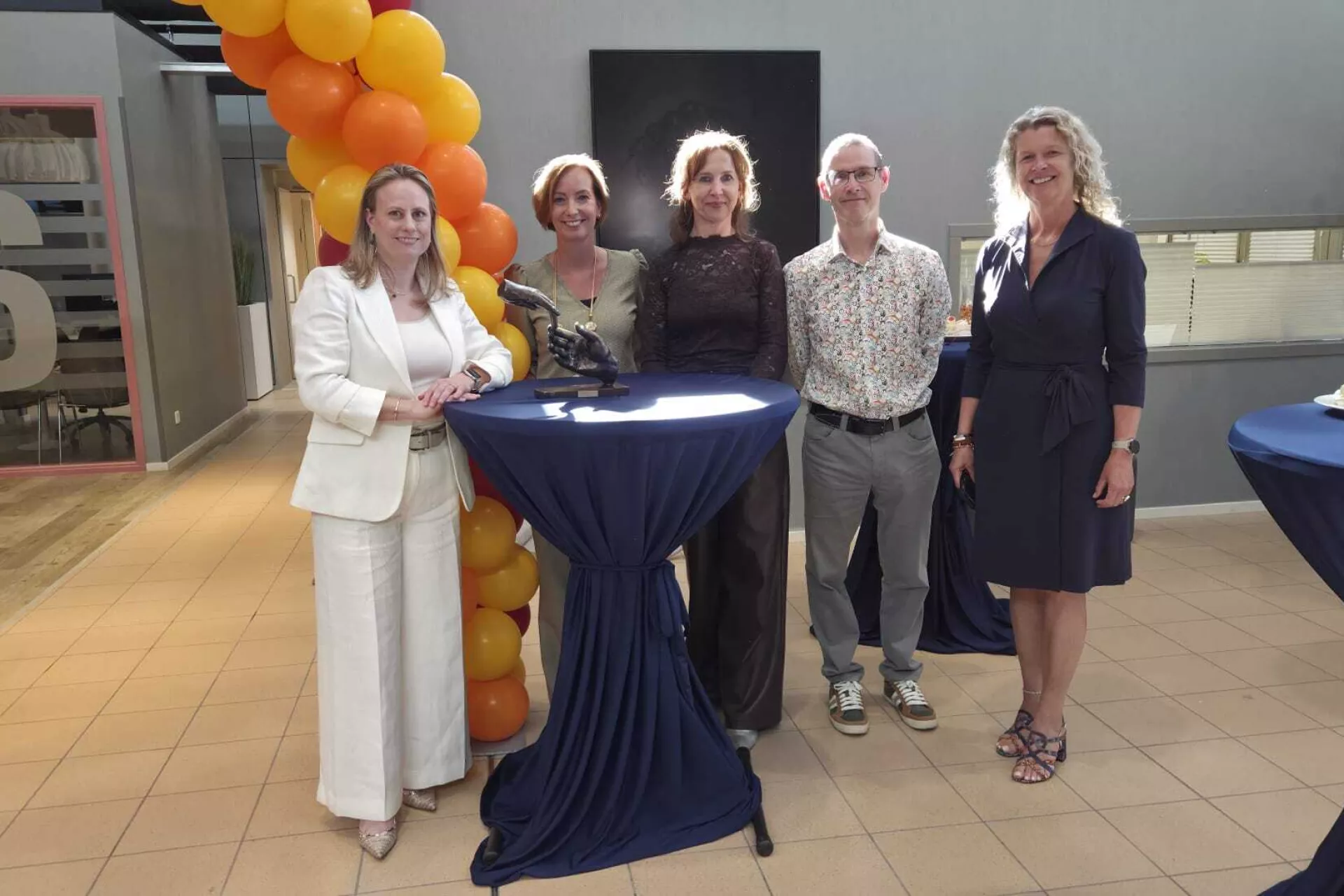 Groepsfoto van vijf mensen bij een sta-tafel met ballondecoratie op de achtergrond.