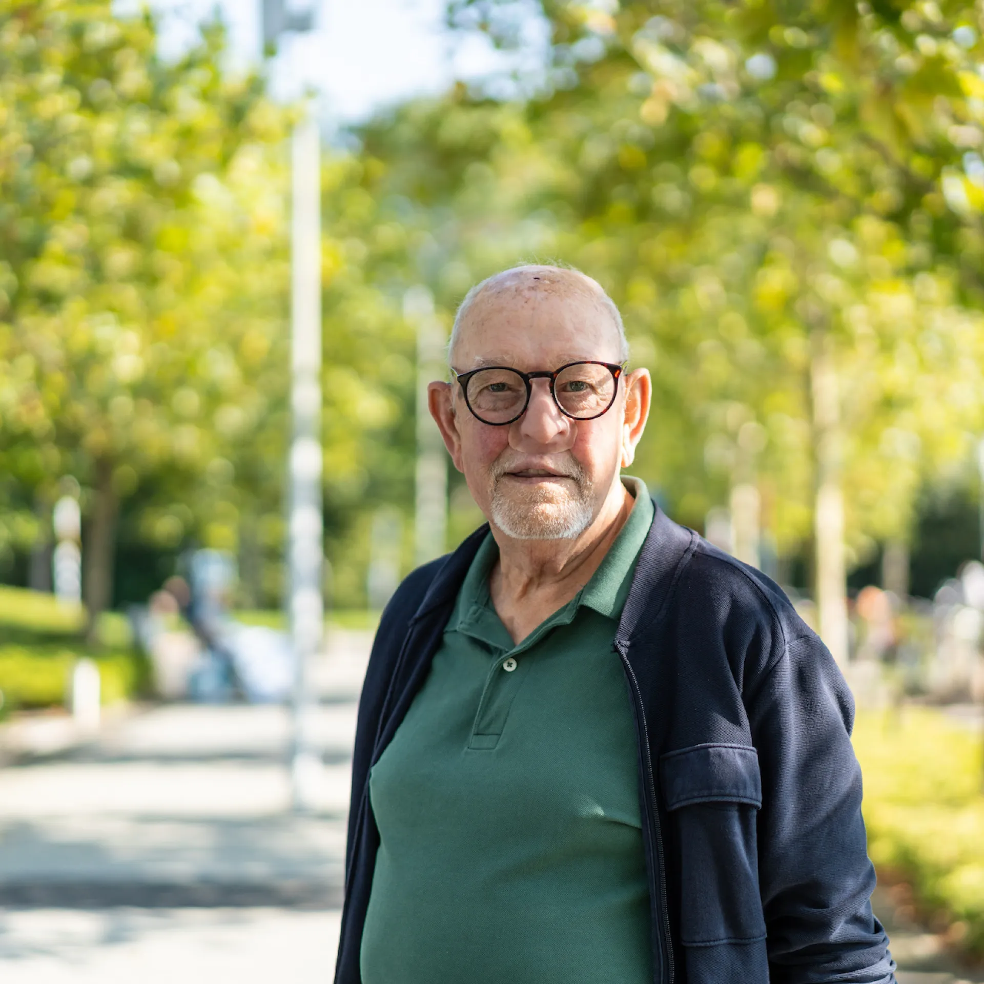 Een oudere man poseert in een zonnig park met bomen op de achtergrond.